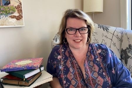 Kelly in a blue and pink patterned shirt, sitting on a couch next to a pile of journals on a table.